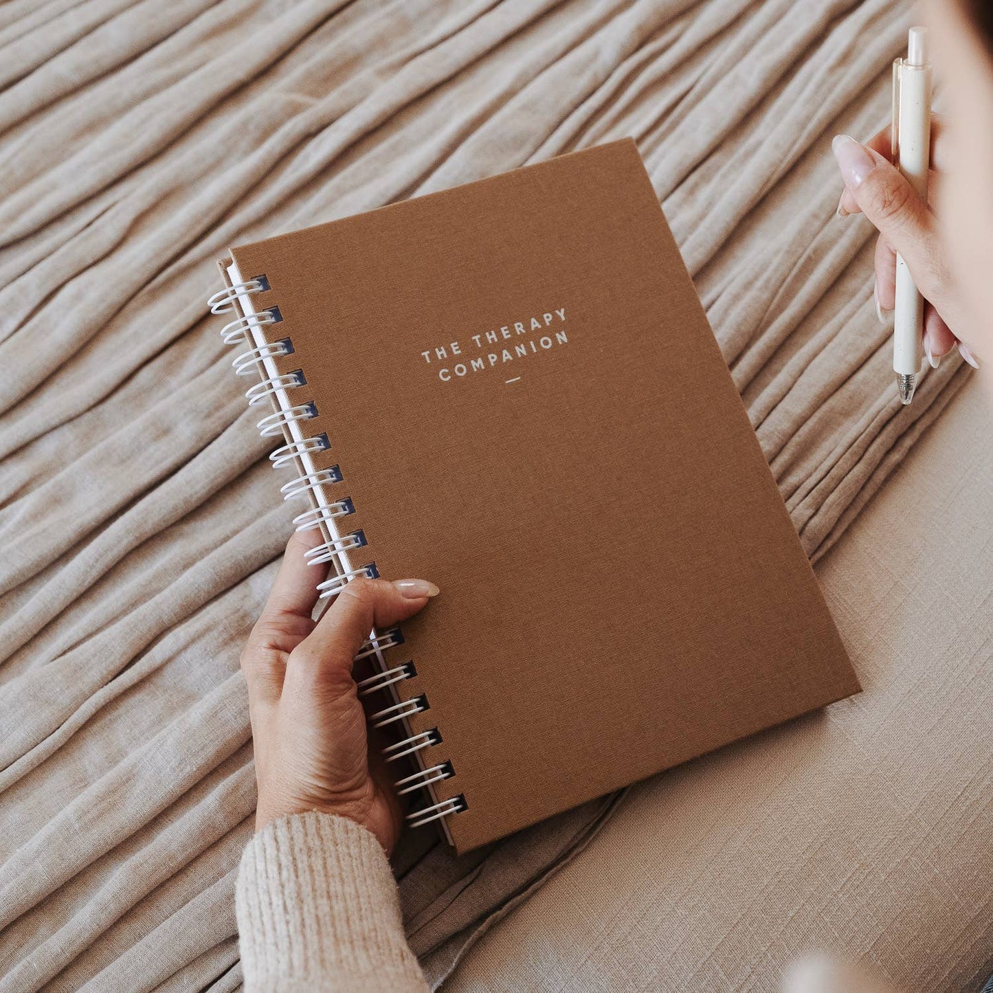 A person holding The Therapy Companion notebook in nutmeg cover while sitting on a bed with a pen in hand.