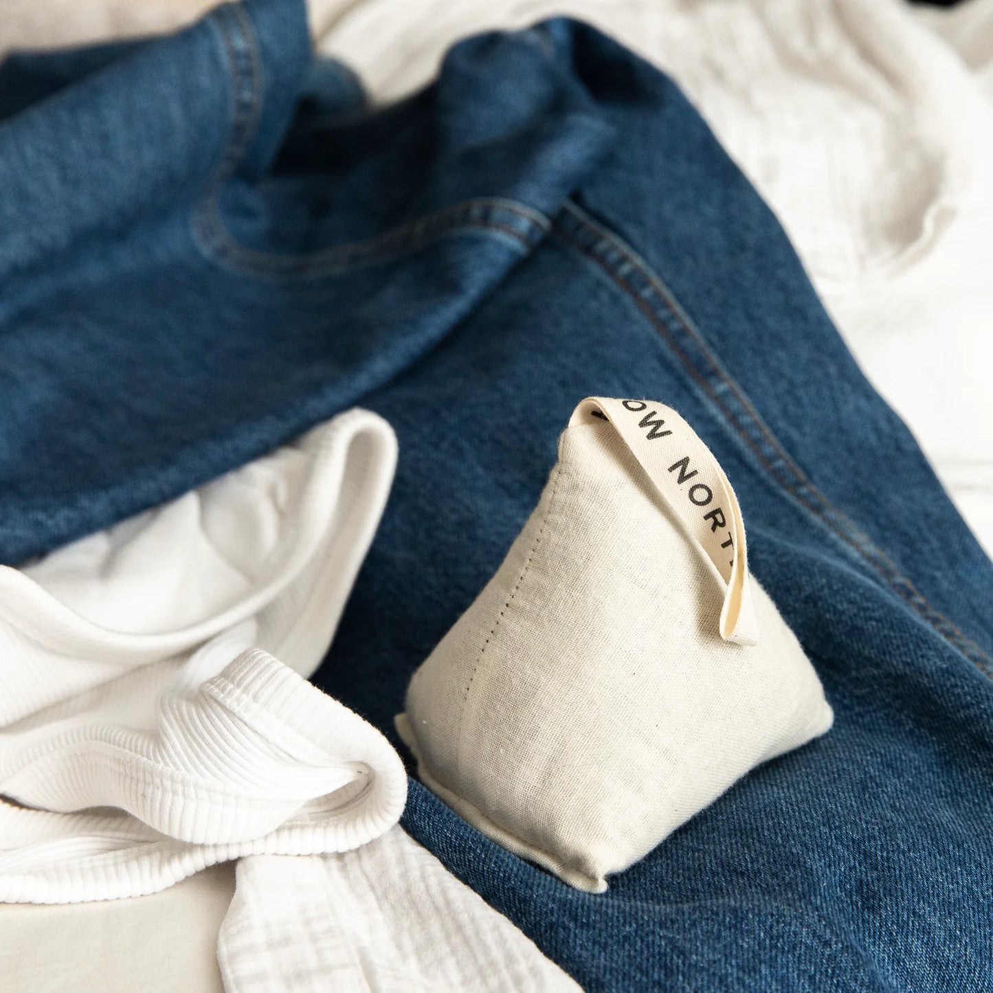 A Slow North Lavender Sachet placed on denim fabric alongside a white ribbed tank and linen, highlighting its simple, natural cotton design.
