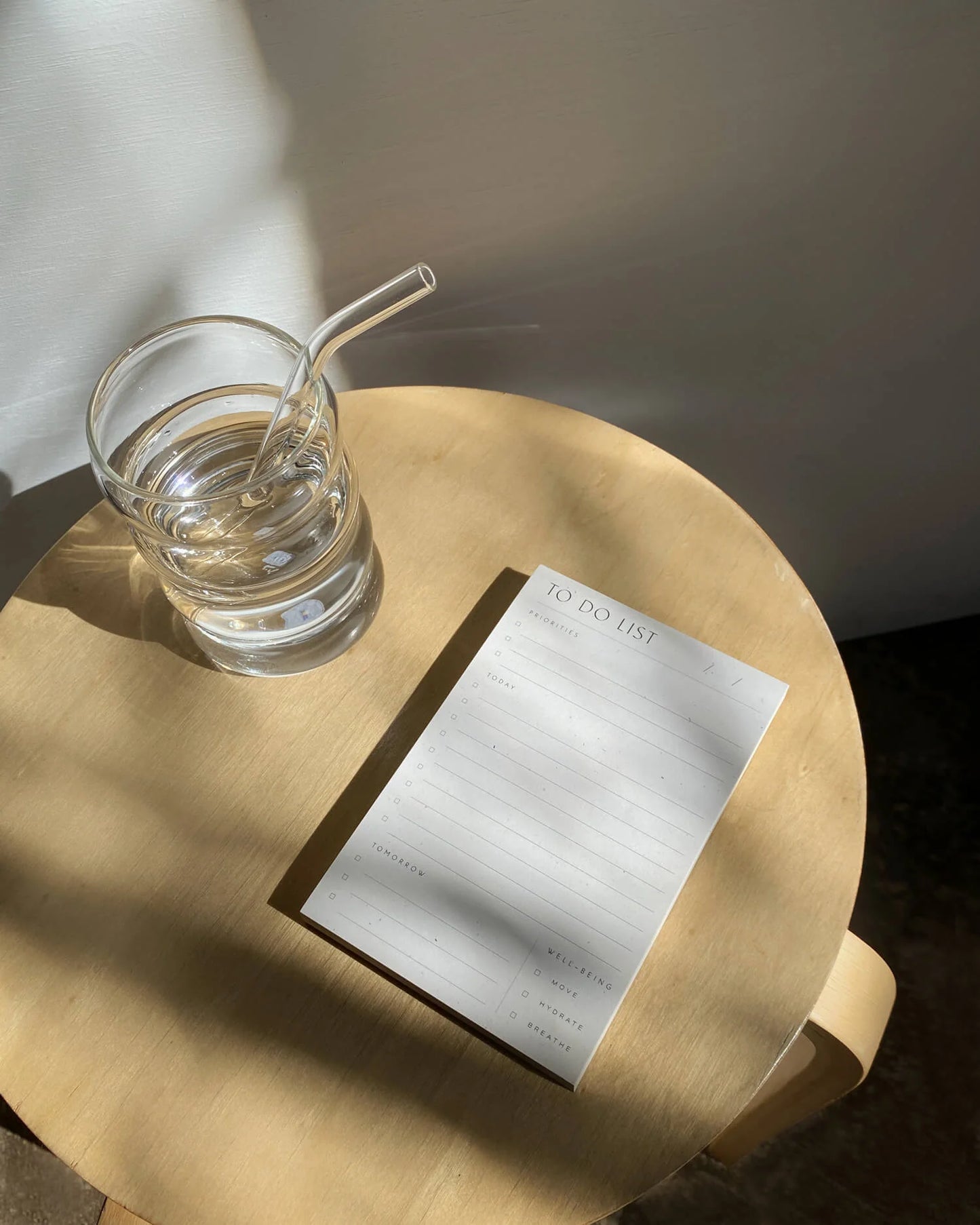 To Do List Pad by Wilde House Paper placed on a round wooden table with a glass of water in sunlight.