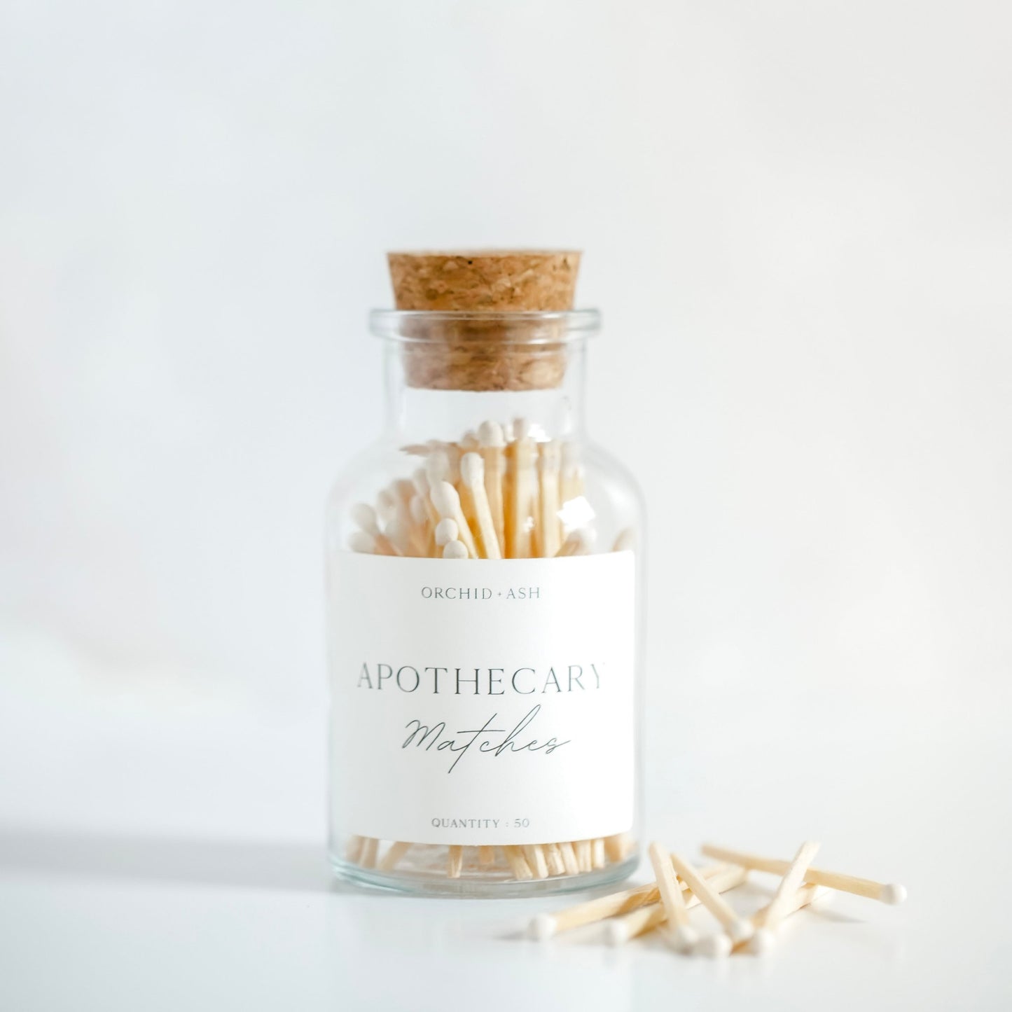 White Apothecary Matches by Orchid & Ash in a clear glass jar with cork top, styled on a white background.