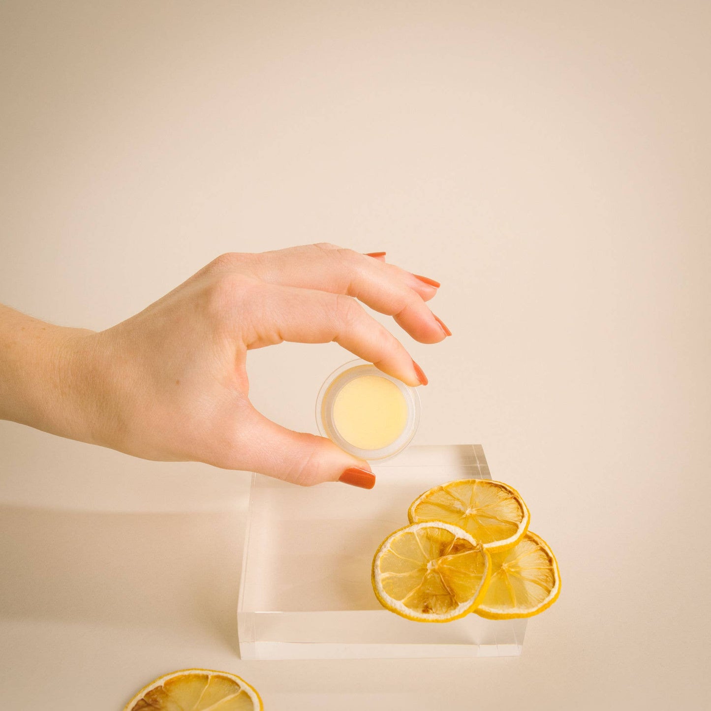 Hand holding an open jar of Sunday Standard Hydrating Lip Butter Salve above a clear block with dried lemon slices, emphasizing natural ingredients.