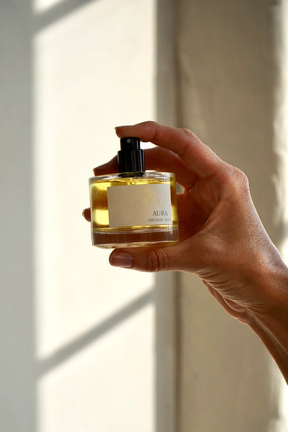 Aura perfume bottle by Orchid & Ash being held by hand with finger on the spritzer with white background.