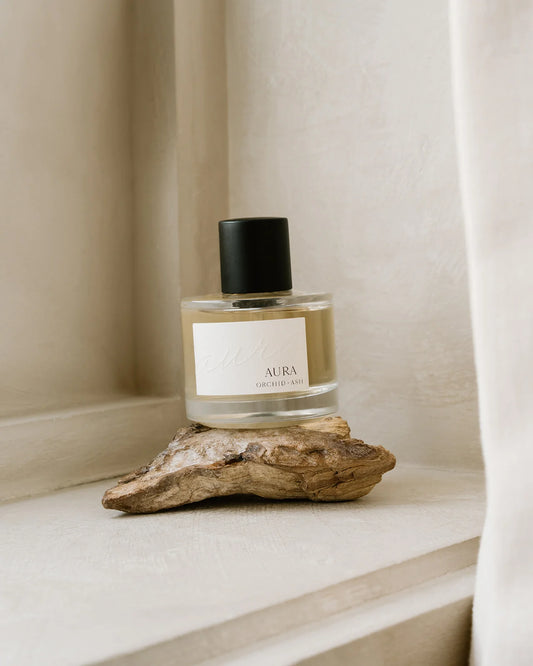 Aura perfume bottle by Orchid & Ash with black cap resting on driftwood with white curtain in background.