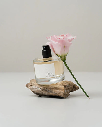Aura fragrance bottle by Orchid & Ash paired with driftwood and a single pink flower on neutral backdrop.