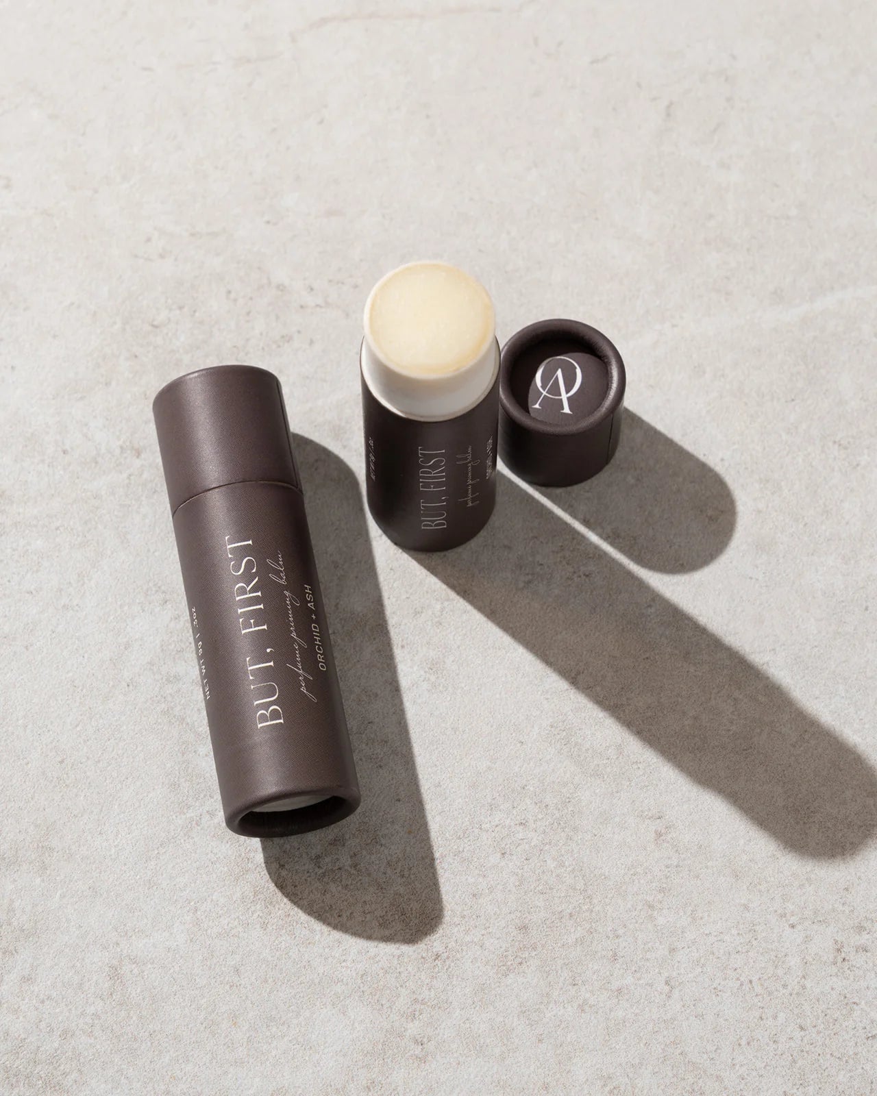 Open tube of But First solid perfume balm by Orchid & Ash casting a soft shadow on a stone surface.