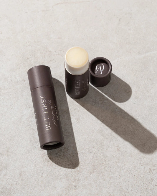 Open tube of But First solid perfume balm by Orchid & Ash casting a soft shadow on a stone surface.