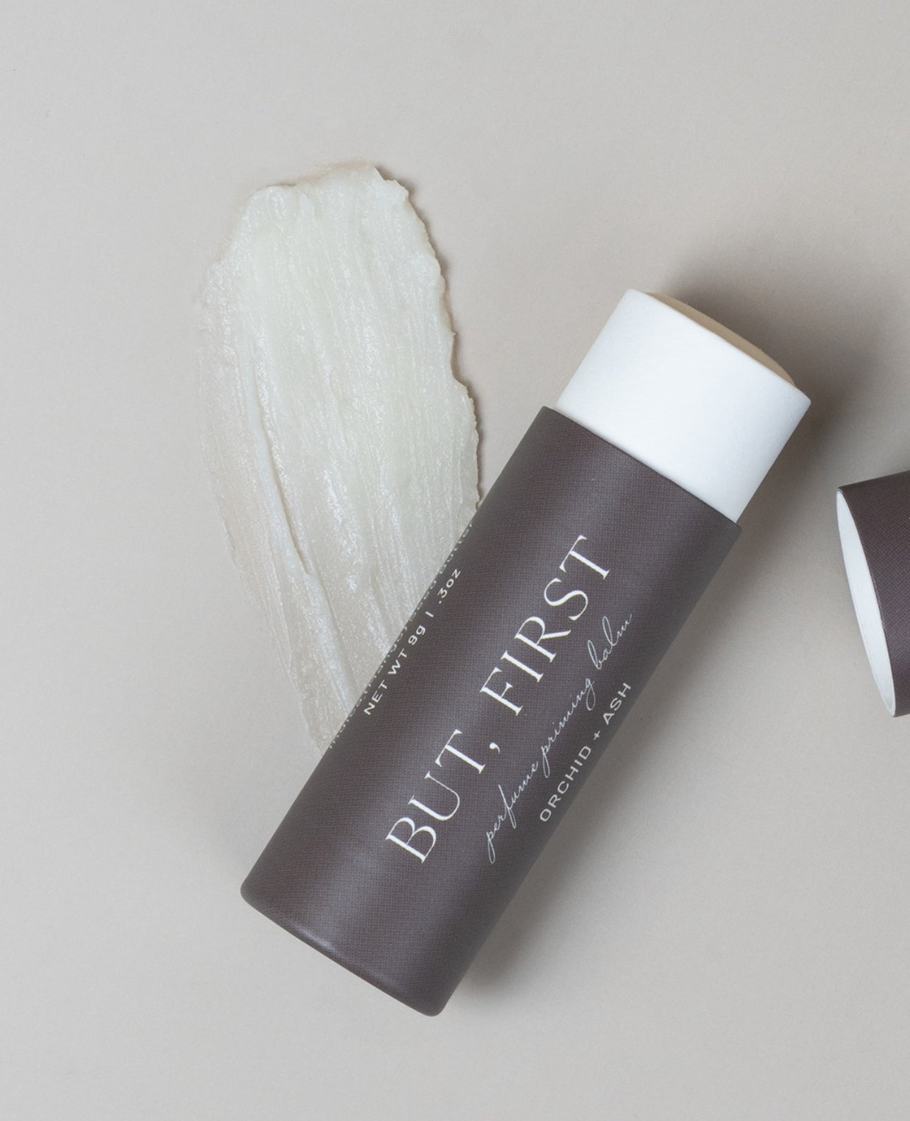 But First solid perfume balm with visible product texture swiped next to the tube on a beige backdrop.