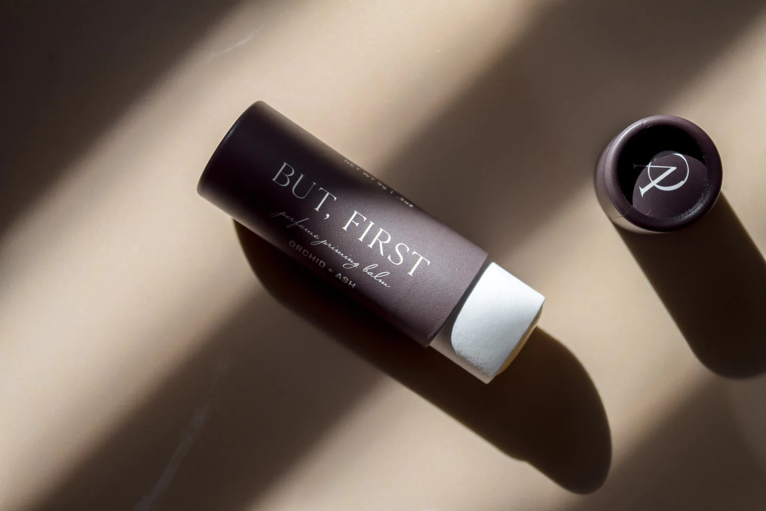 Top-down view of But First perfume balm and cap by Orchid & Ash on a soft neutral background.