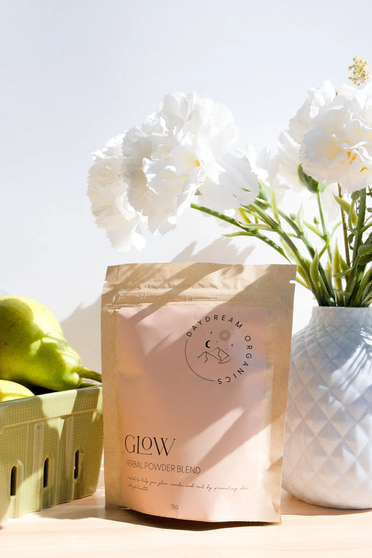 Daydream Organics Glow Herbal Powder Blend resting on a table next to pears and a vase of flowers.