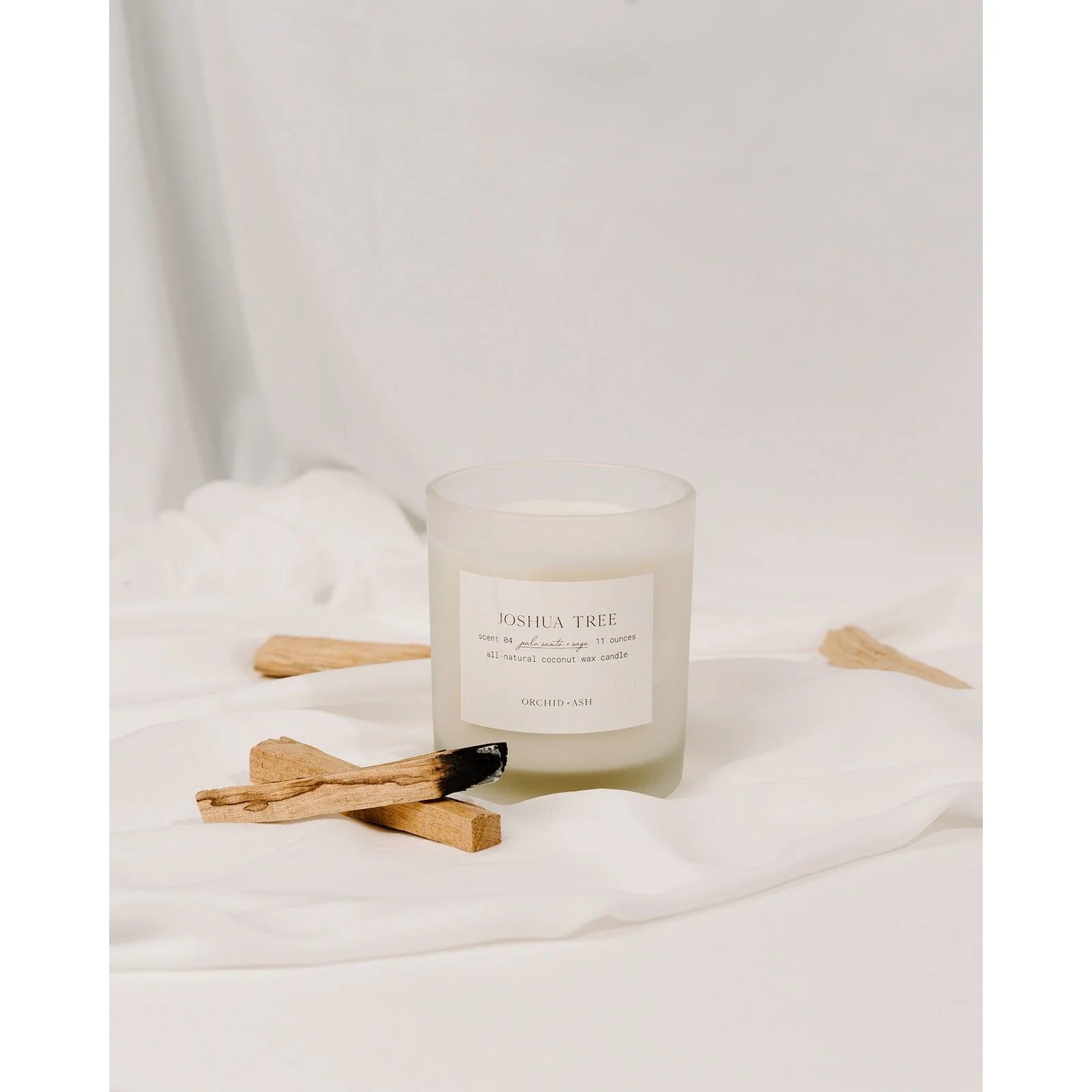 Minimalist shot of Orchid & Ash Joshua Tree candle on soft fabric with palo santo pieces.