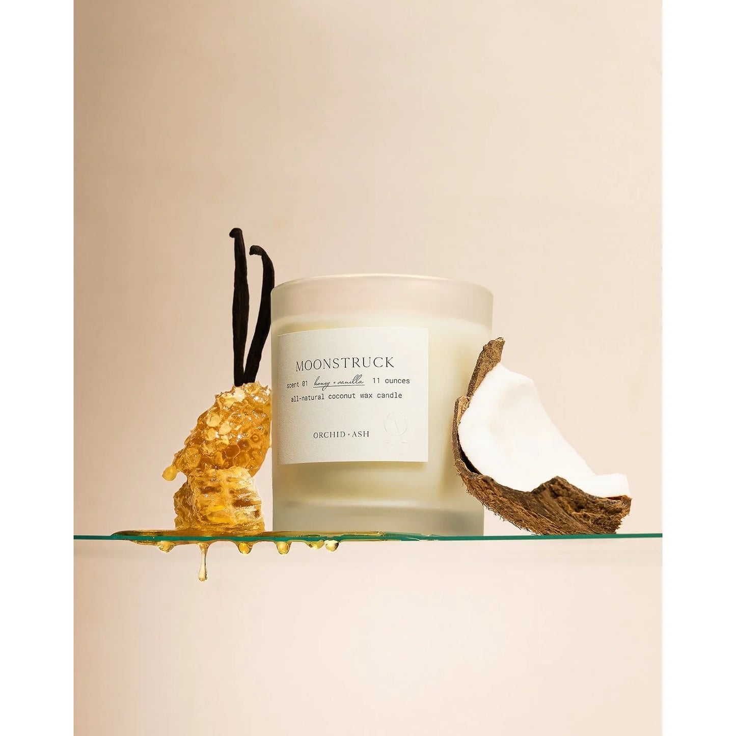 Frosted Moonstruck candle jar by Orchid & Ash next to a honeycomb, vanilla bean, and halved coconut on a glass shelf.