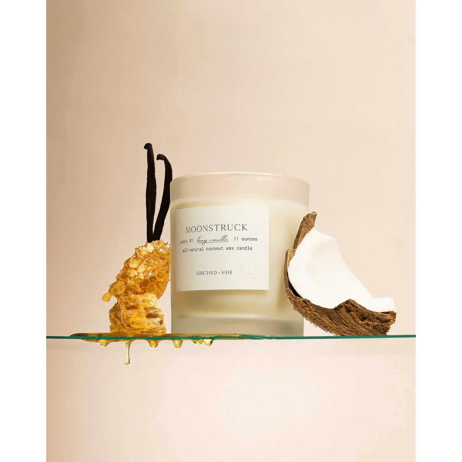 Frosted Moonstruck candle jar by Orchid & Ash next to a honeycomb, vanilla bean, and halved coconut on a glass shelf.