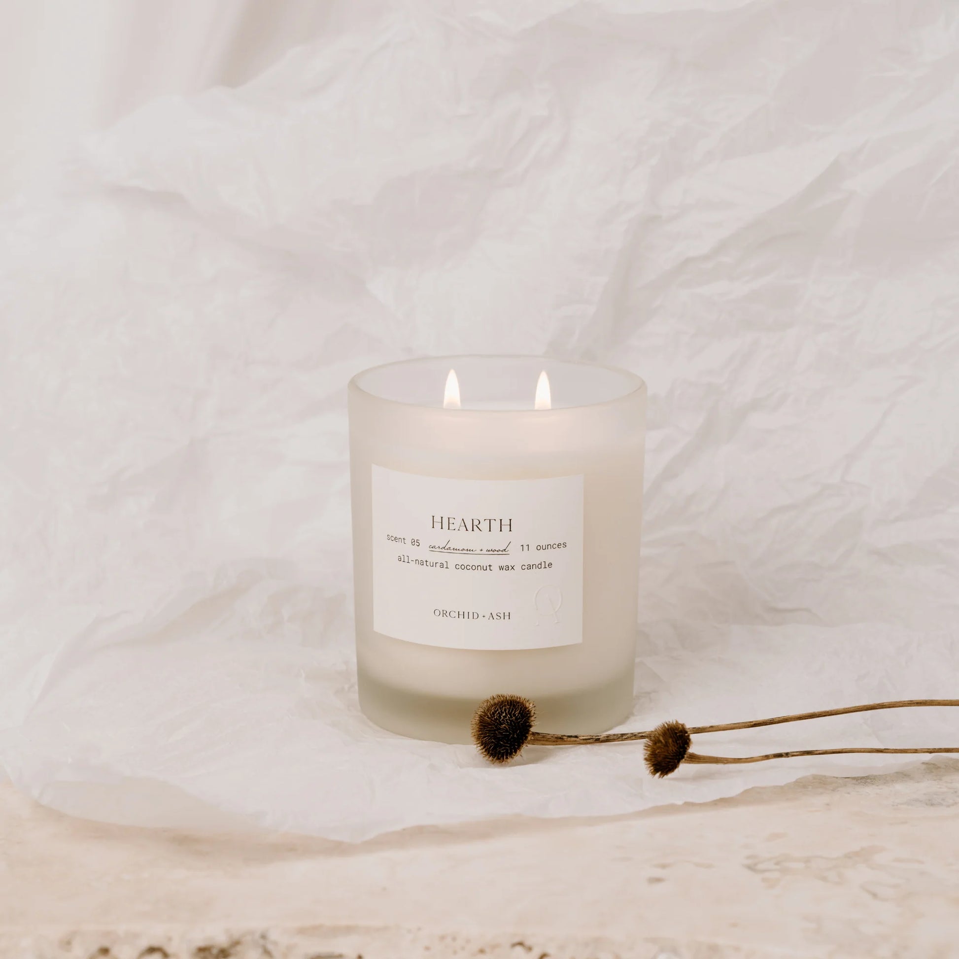Hearth candle by Orchid & Ash glowing on crumpled white fabric with dried flowers.