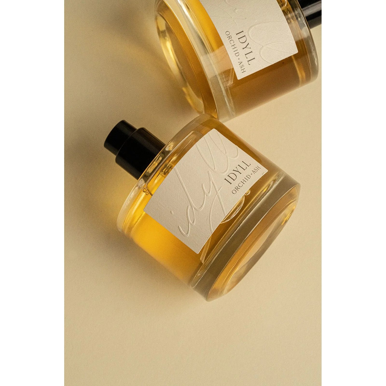 Two Idyll perfume bottles by Orchid & Ash lying on a warm beige background with golden light.