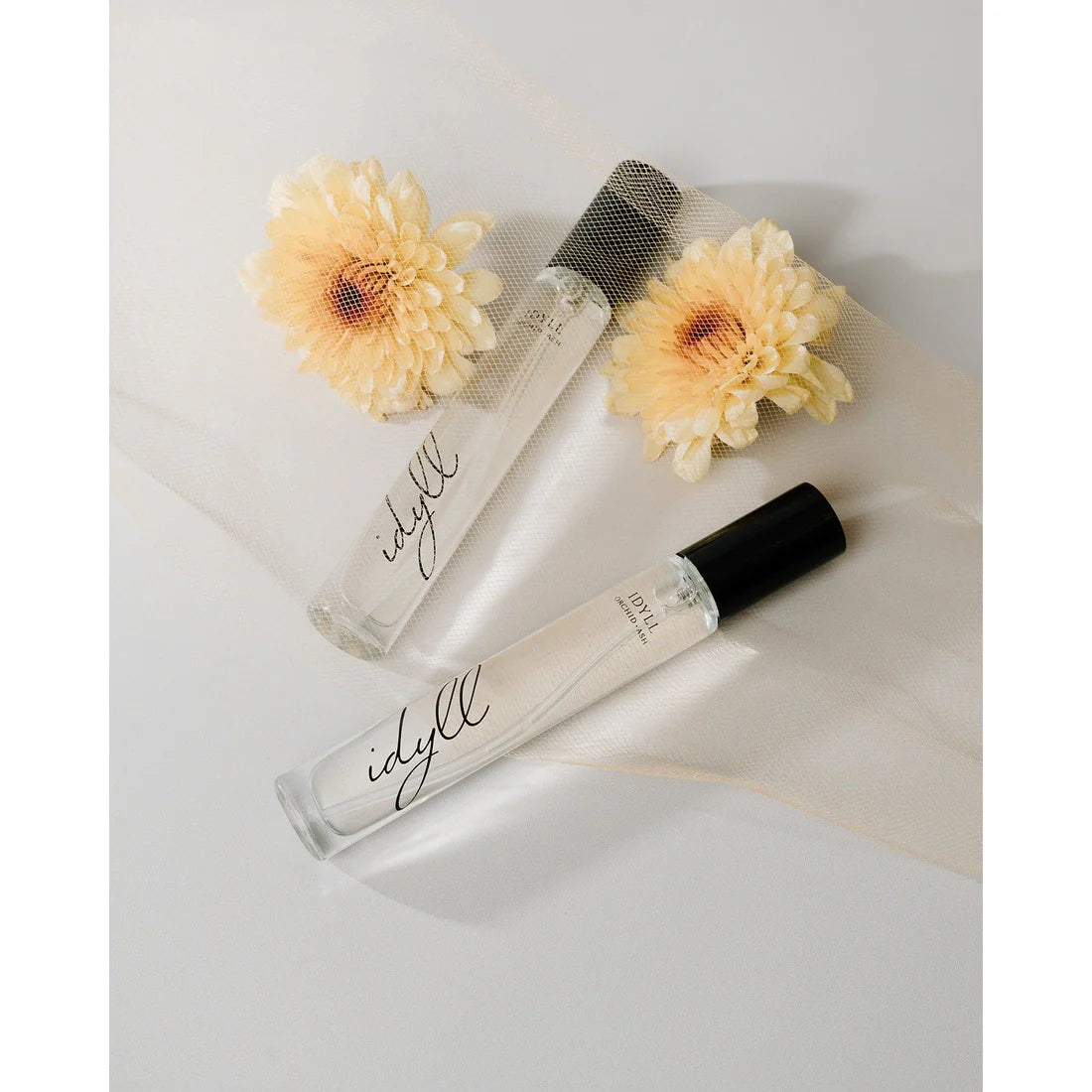 Idyll perfume travel bottles by Orchid & Ash placed on sheer fabric alongside soft yellow daisies.