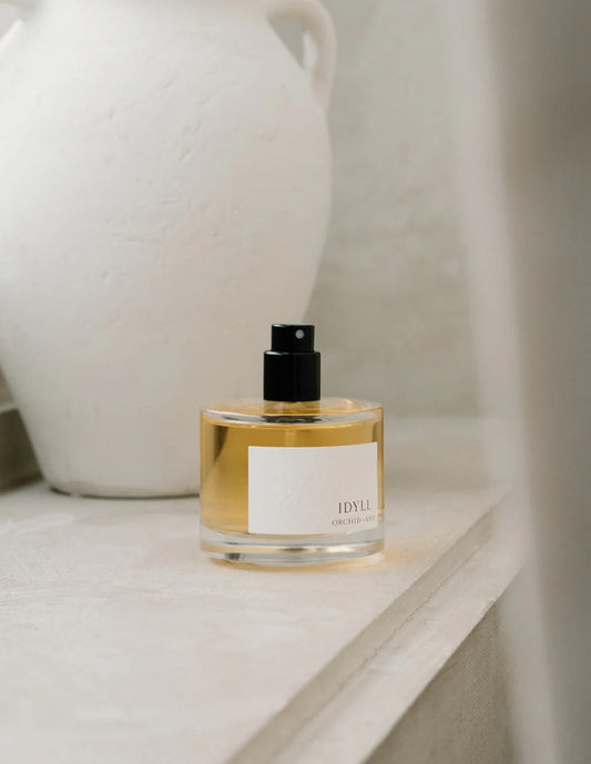 Idyll eau de parfum bottle by Orchid & Ash resting on the edge of a stone bathtub beside a white ceramic vase.