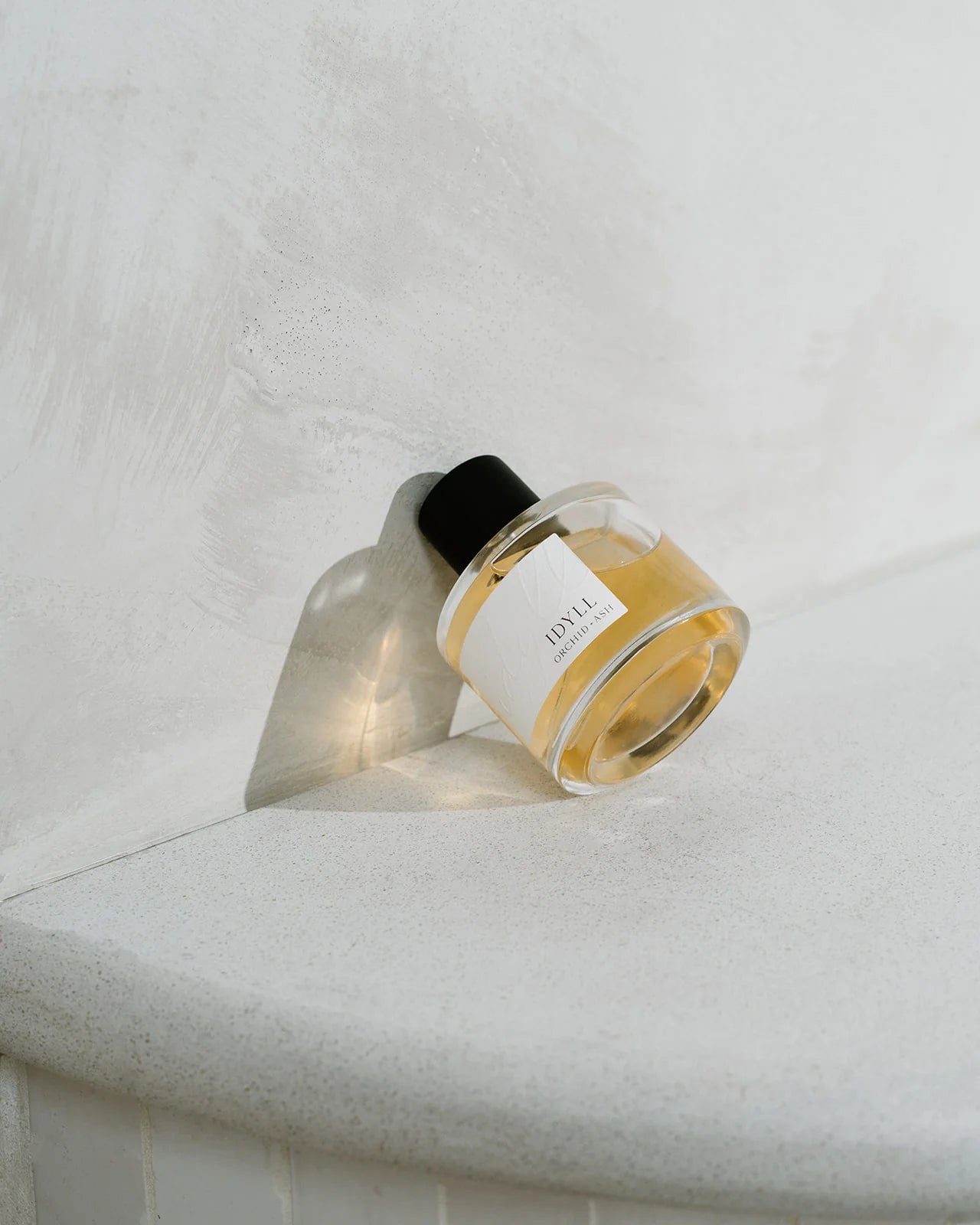 Idyll perfume bottle by Orchid & Ash casting a soft shadow on a curved, minimal plaster wall.