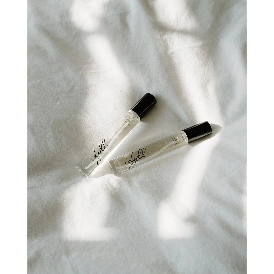 Two Idyll travel spray perfumes by Orchid & Ash photographed side-by-side in natural light on linen fabric.