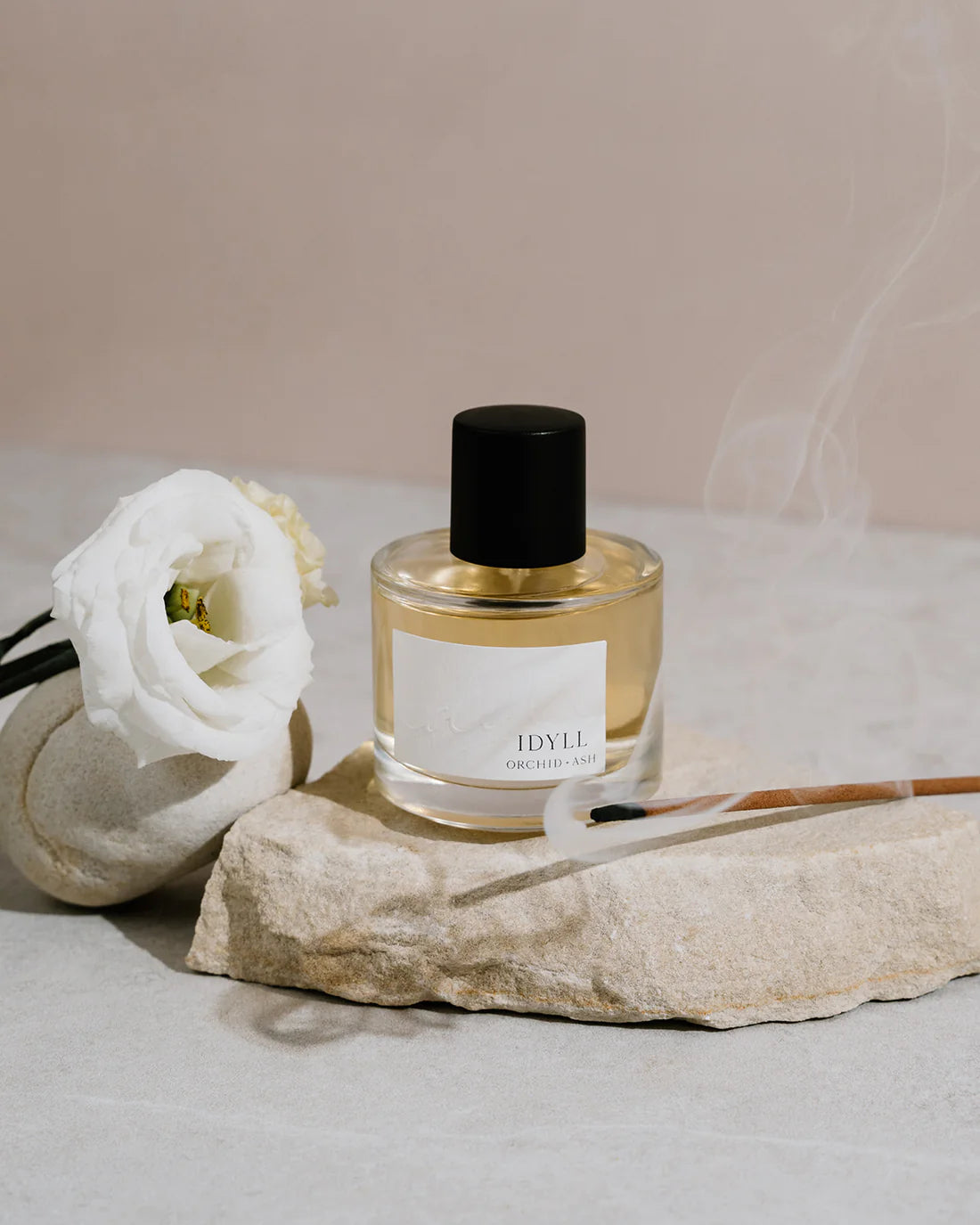 Idyll perfume by Orchid & Ash styled with incense smoke and a white flower on a stone surface.