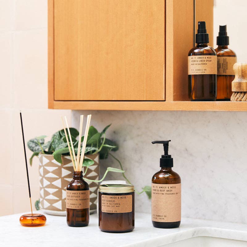 Full P.F. Candle Co. Amber & Moss collection styled in bathroom setting with soap, incense, and diffuser by a ceramic pot with greenery.