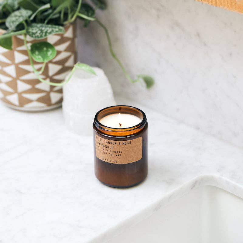 P.F. Candle Co. Amber & Moss Candle lit on white bathroom counter next to ceramic vase and greenery.