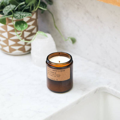 P.F. Candle Co. Amber & Moss Candle lit on white bathroom counter next to ceramic vase and greenery.