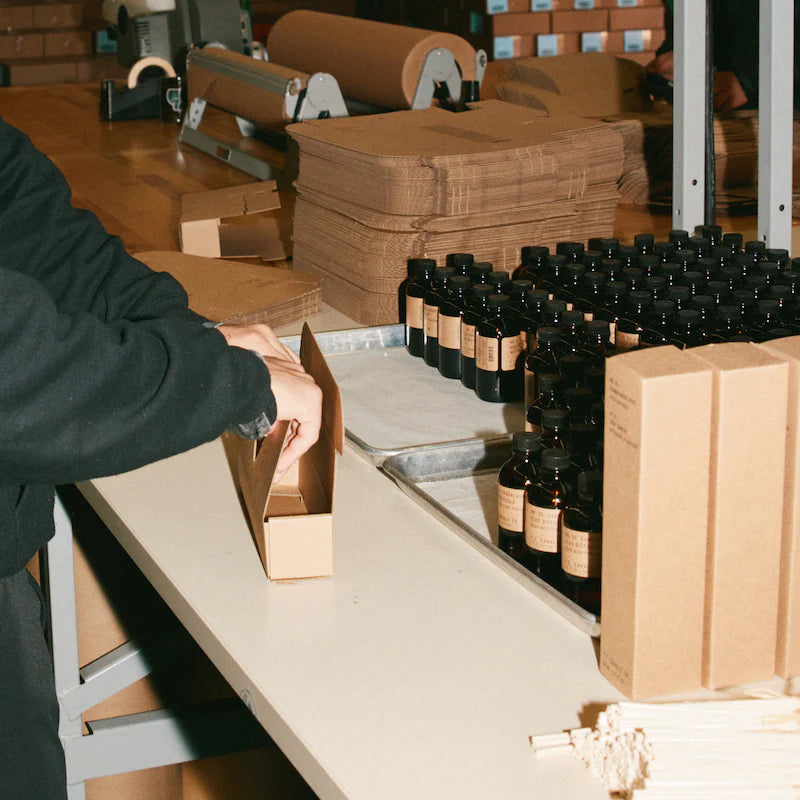 P.F. Candle Co. staff packaging reed diffusers on a production line with amber bottles and kraft boxes.