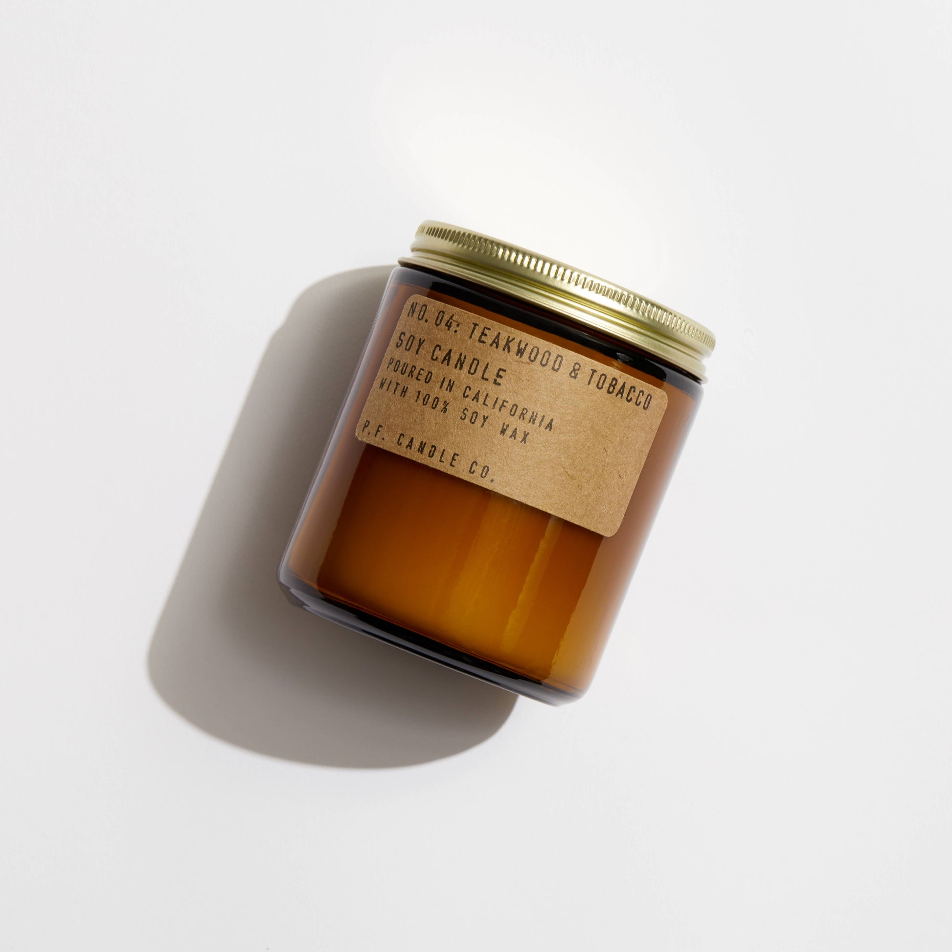 Teakwood & Tobacco soy candle by P.F. Candle Co. laying on white background, casting soft shadow.