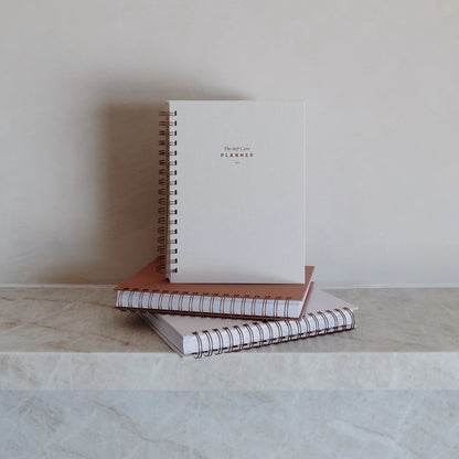 The Self Care Planner in sand and dusty rose stacked on top of other planners on a stone shelf.