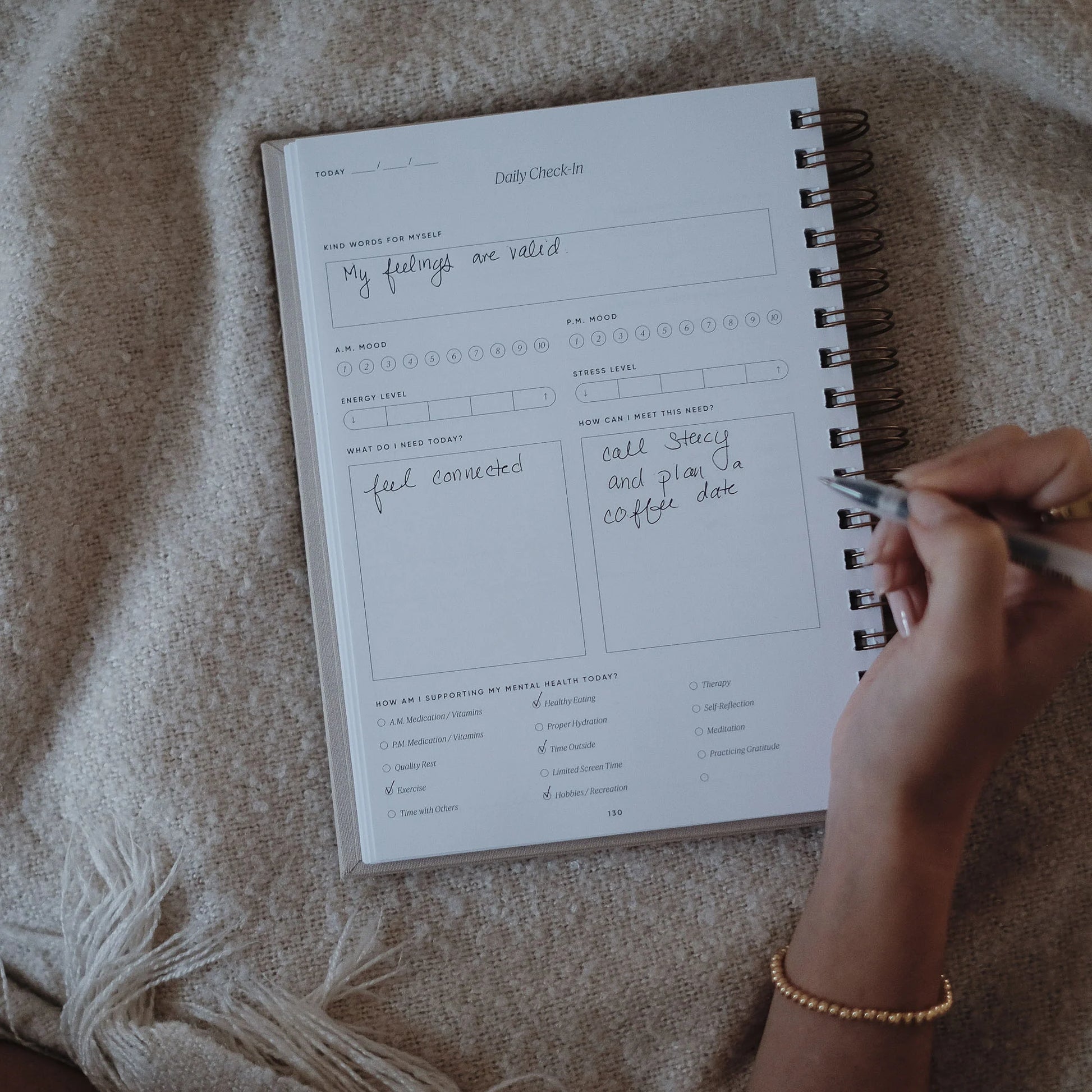 Hands writing in the daily check-in section of The Inner Work Journal, resting on a cozy blanket.