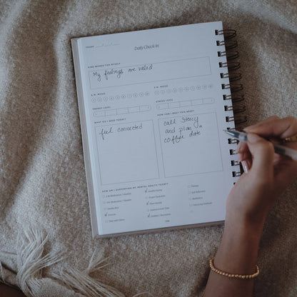 Hands writing in the daily check-in section of The Inner Work Journal, resting on a cozy blanket.