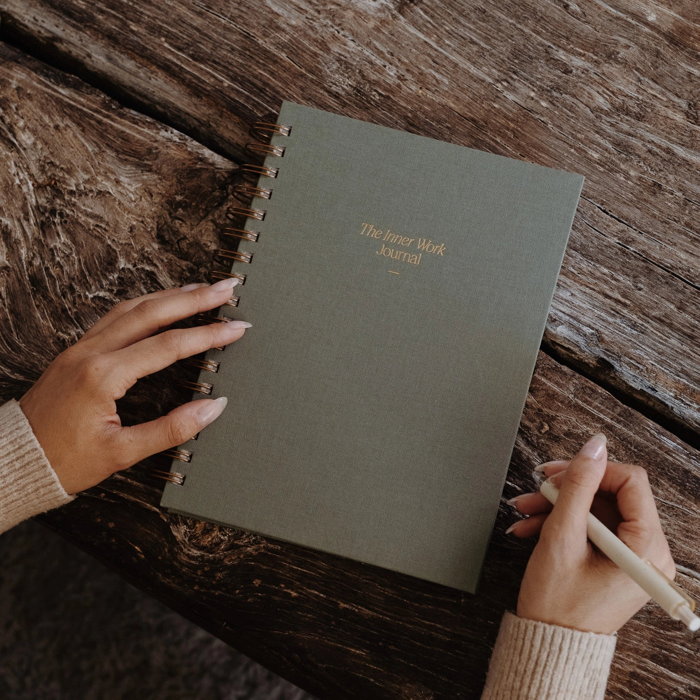The Inner Work Journal in sage placed on a rustic wooden table, with a person holding a pen ready to write.
