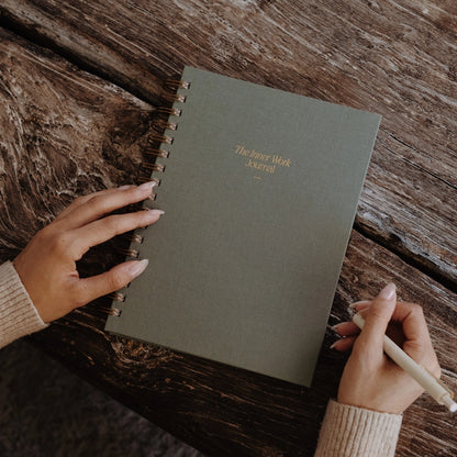 The Inner Work Journal in sage placed on a rustic wooden table, with a person holding a pen ready to write.
