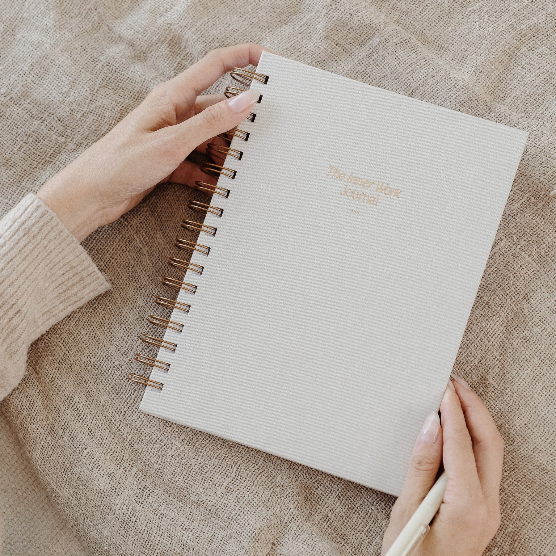Hands gently holding The Inner Work Journal in sand, placed on a linen fabric surface.