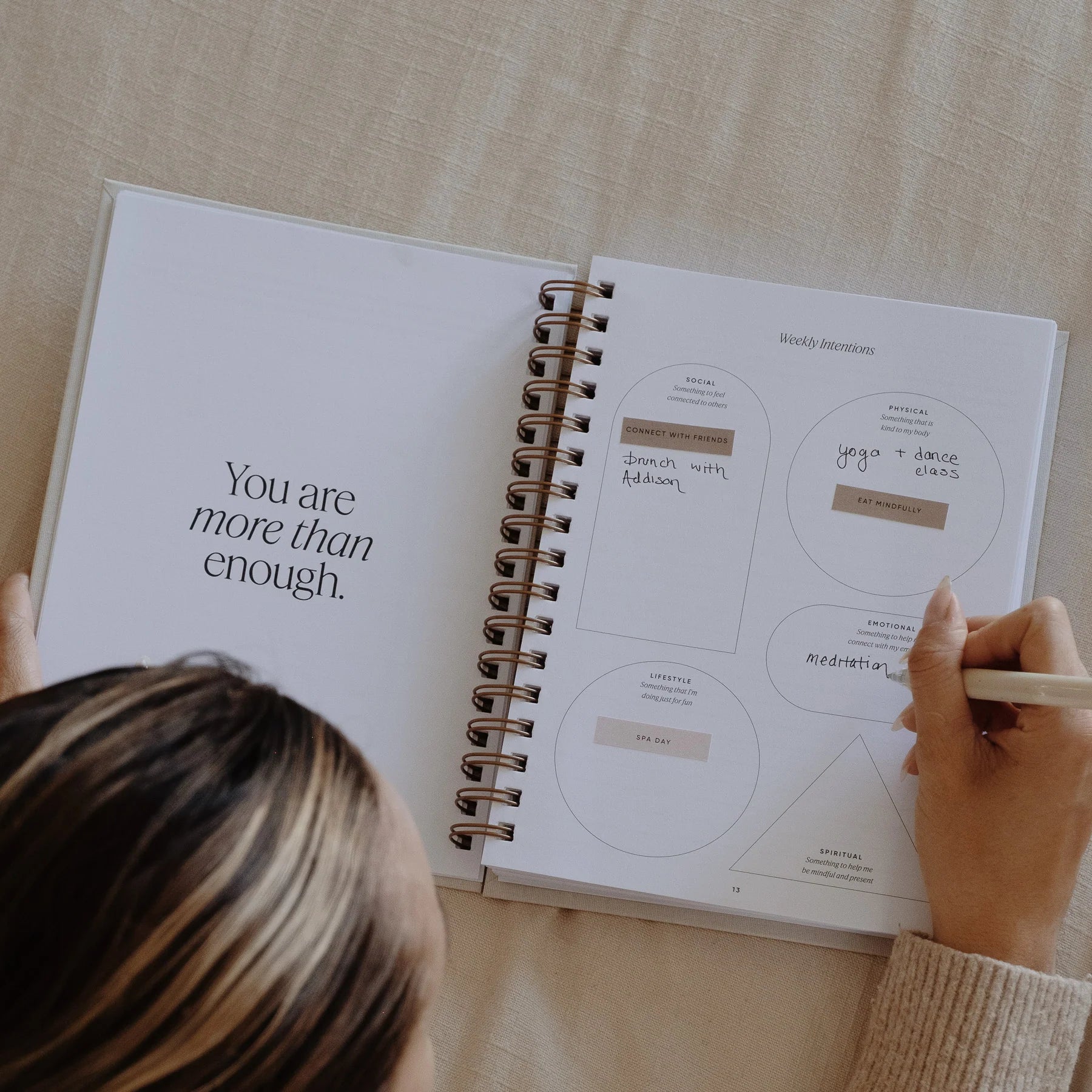 The Inner Work Journal open to the weekly intentions page with a motivational quote "You are more than enough," on the left page and a person writing down goals for upcoming week with stickers.