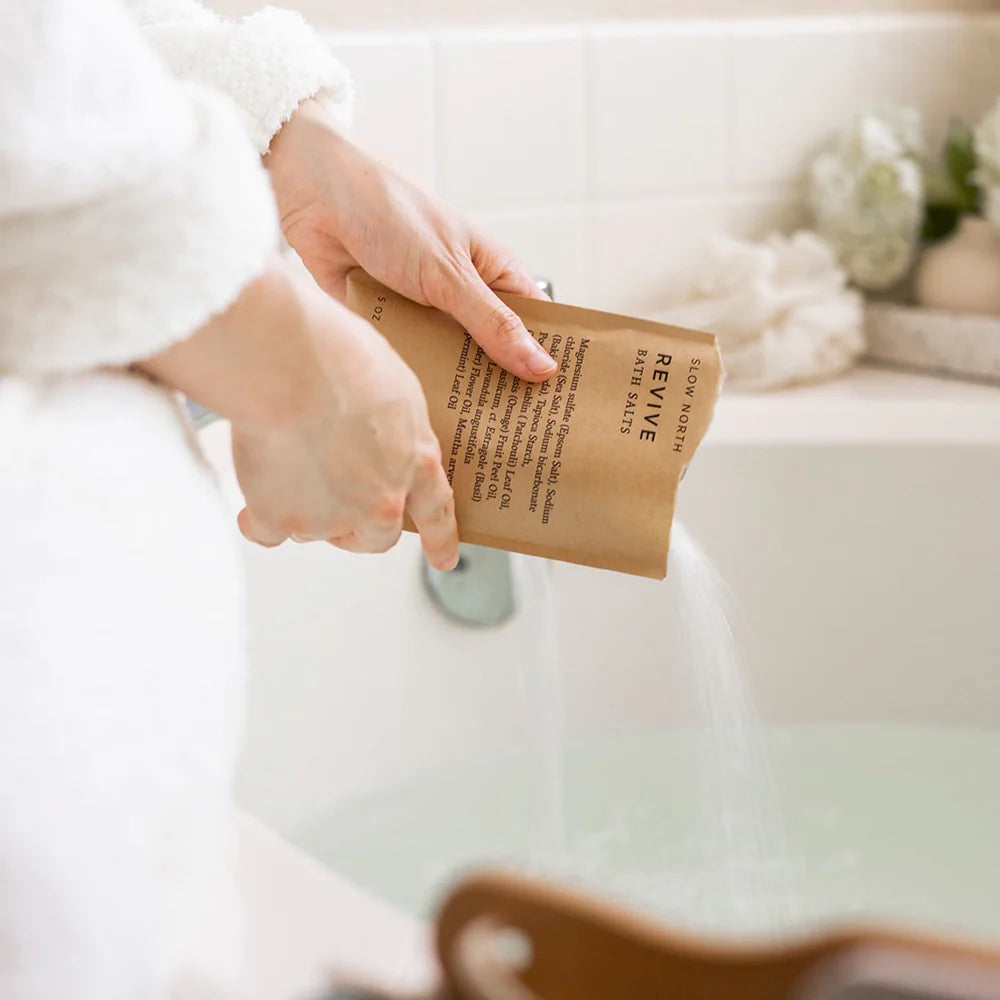 A person in a white robe pours Slow North Revive Bath Salts into a bathtub, creating a serene spa-like atmosphere.