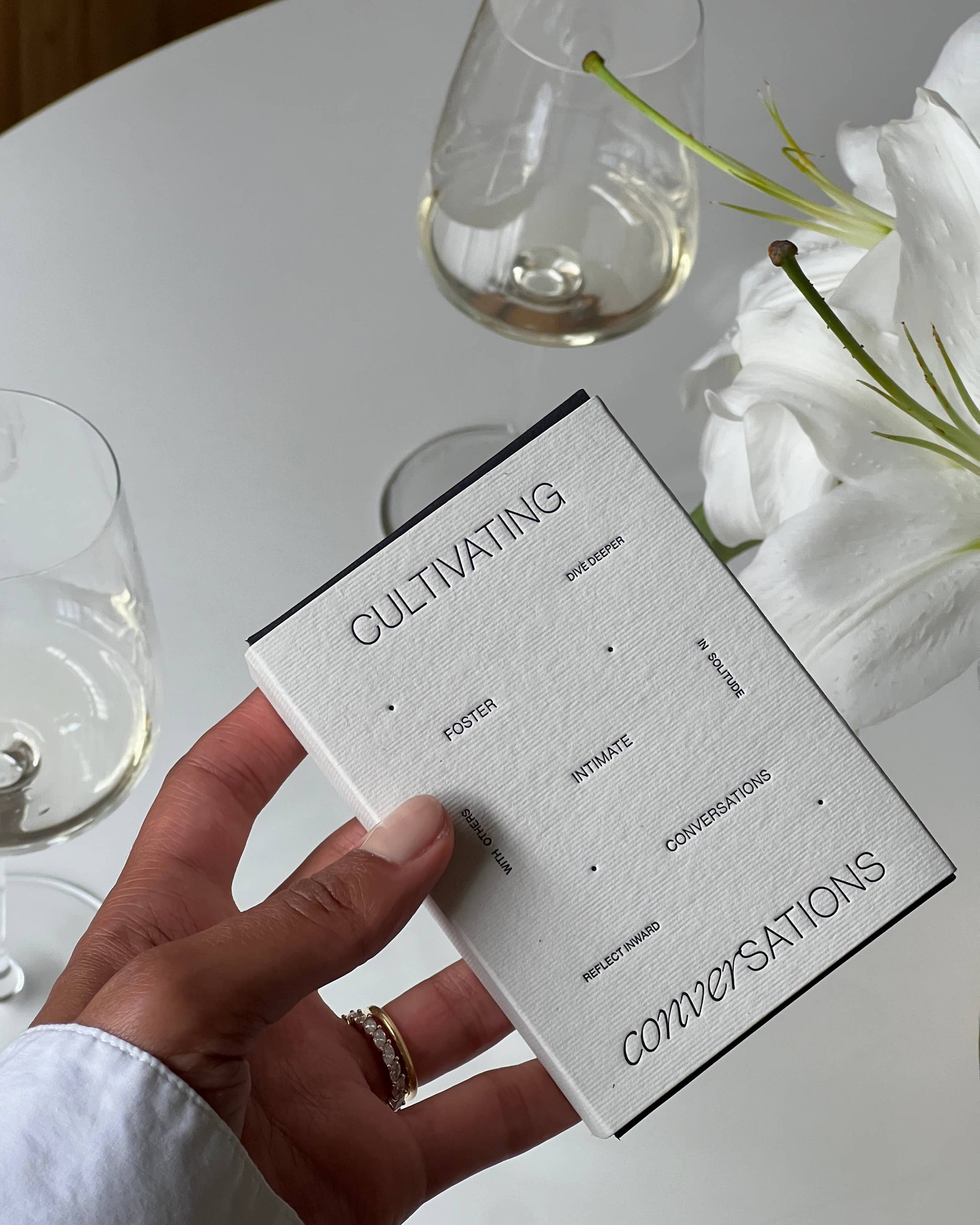 Hand holding the Wilde House Paper Cultivating Conversations Card Deck box next to glasses of white wine and white flowers.