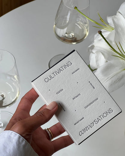 Hand holding the Wilde House Paper Cultivating Conversations Card Deck box next to glasses of white wine and white flowers.