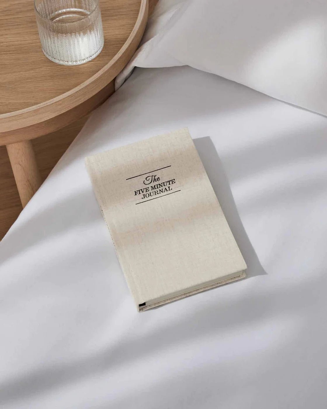 The Five Minute Journal styled on a bedside table and bed, promoting mindful morning and evening routines.