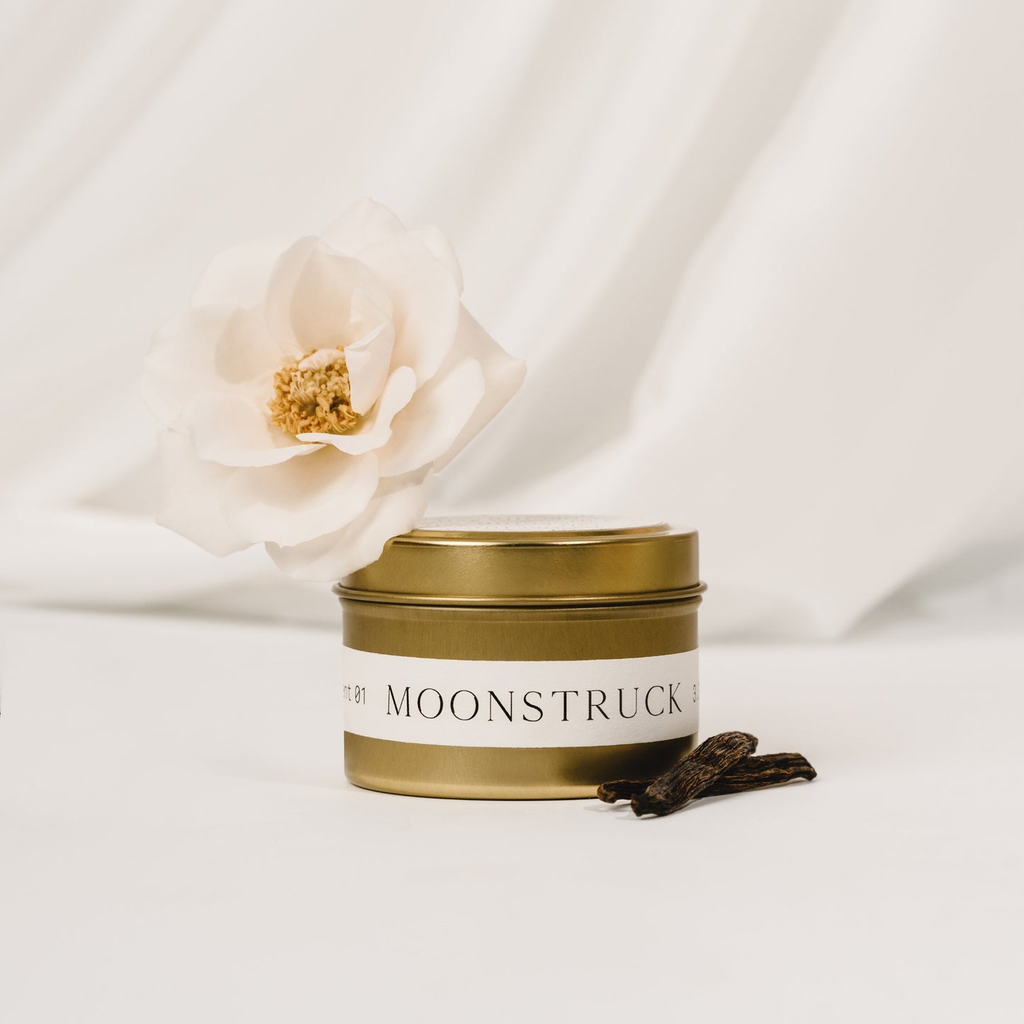 Moonstruck candle tin by Orchid & Ash surrounded by vanilla beans and soft white flower on draped fabric.