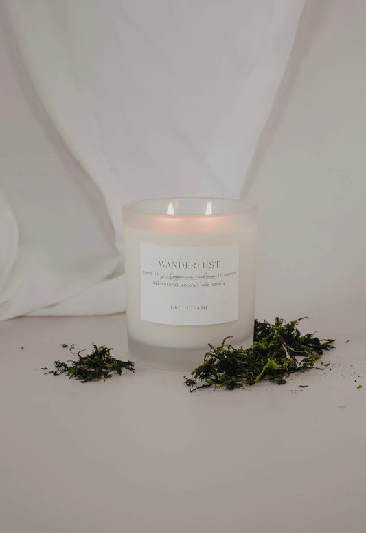 Wanderlust candle by Orchid & Ash surrounded by scattered oakmoss on a soft fabric background.