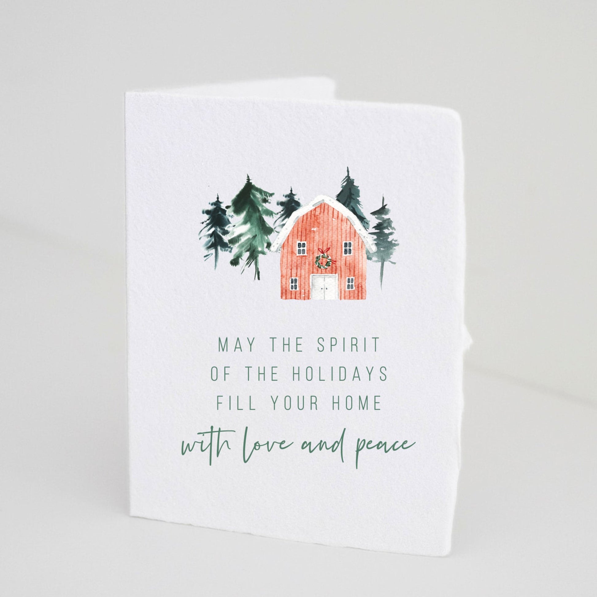 Front of Paper Baristas holiday card featuring an illustration of a cozy cabin and evergreen trees with the message “May the spirit of the holidays fill your home with love and peace.
