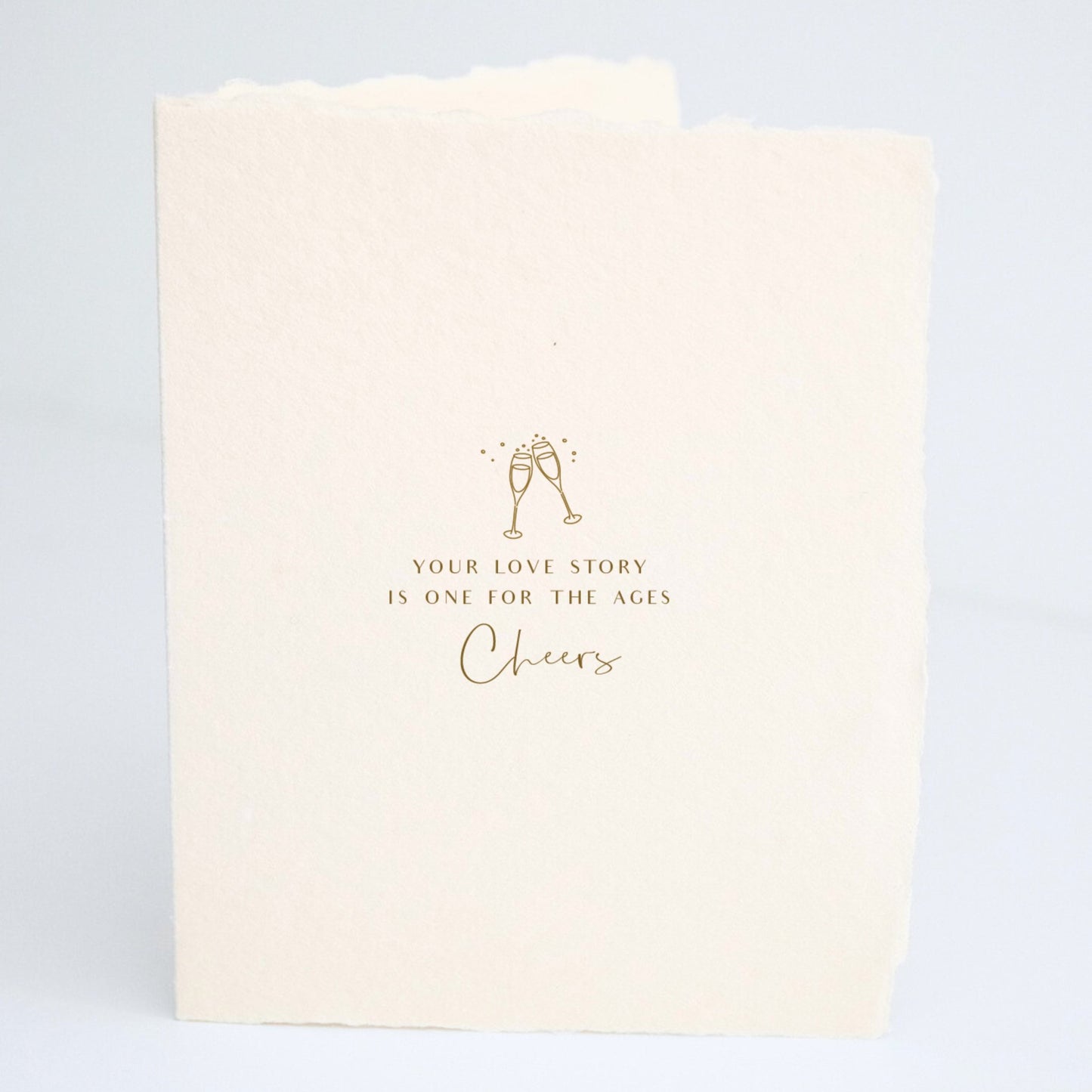 Front of Paper Baristas wedding card with champagne glasses illustration and the words “Your love story is one for the ages. Cheers" in gold foil.