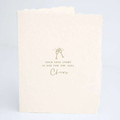 Front of Paper Baristas wedding card with champagne glasses illustration and the words “Your love story is one for the ages. Cheers" in gold foil.