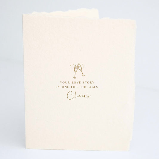 Front of Paper Baristas wedding card with champagne glasses illustration and the words “Your love story is one for the ages. Cheers" in gold foil.