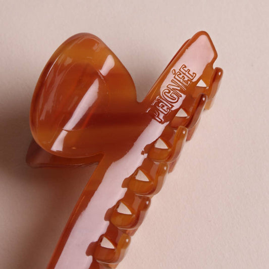 Close-up of amber caramel colored French hair claw clip with Peignée Paris engraved logo branding, sculpted curve and glossy finish.
