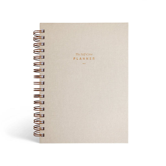 The Self Care Planner Daily Edition in sand, closed and upright with gold foil lettering on the cover.