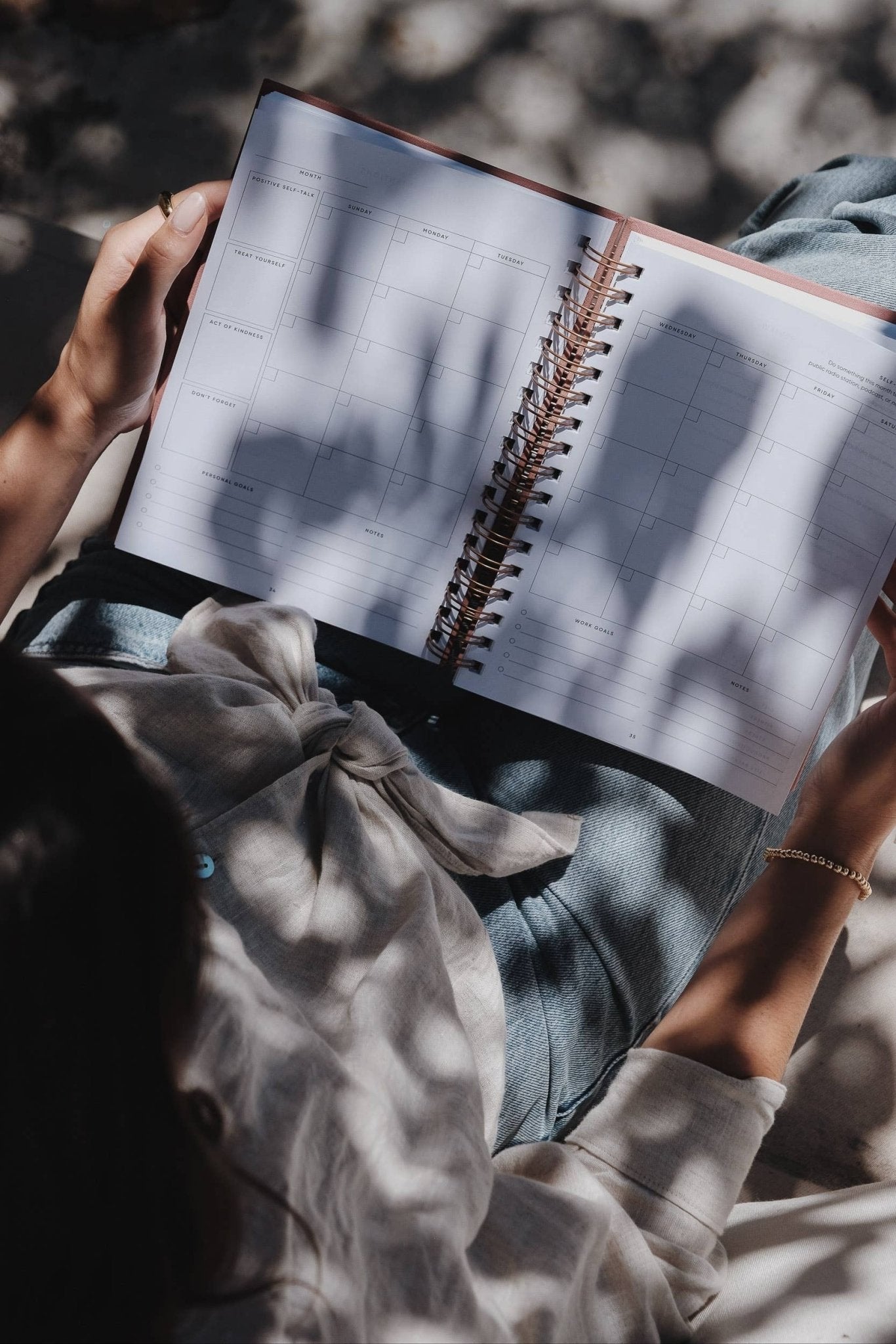 Person sitting outside holding The Self Care Planner on their lap, open to the monthly overview spread under soft sunlight.