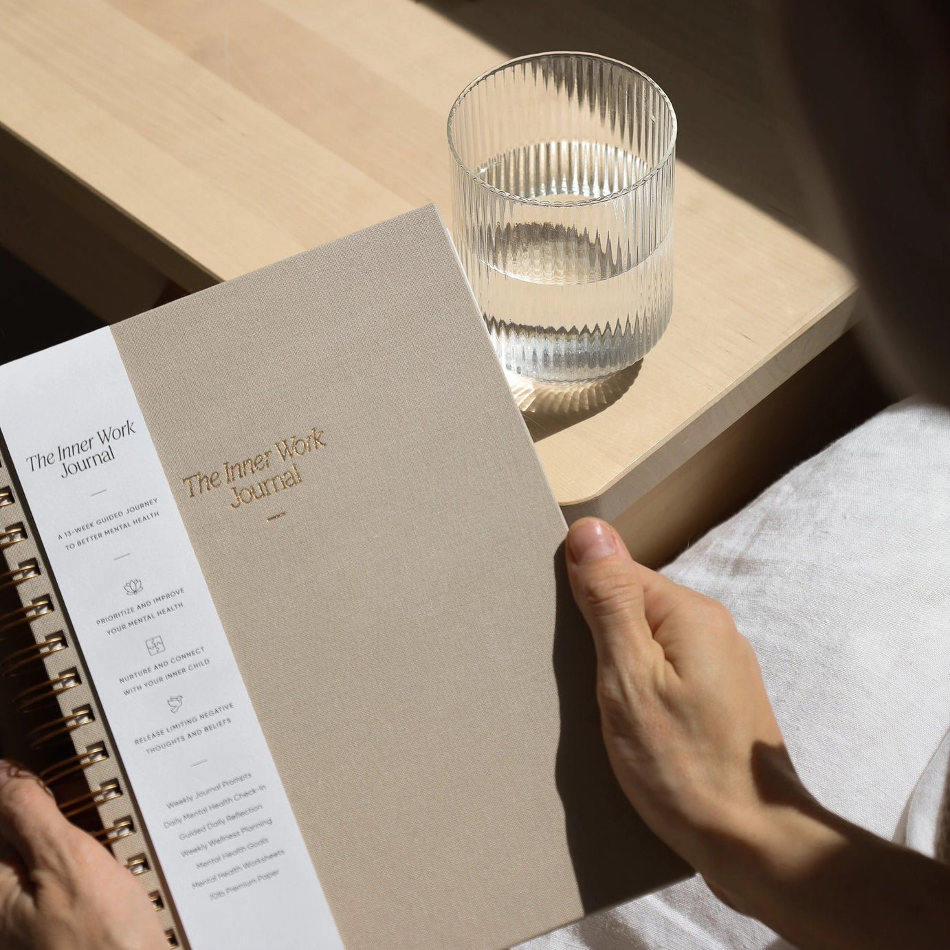 The Inner Work Journal in a sand beige color packaged with belly band, being held near a glass of water on a natural wooden table, bathed in natural sunlight.