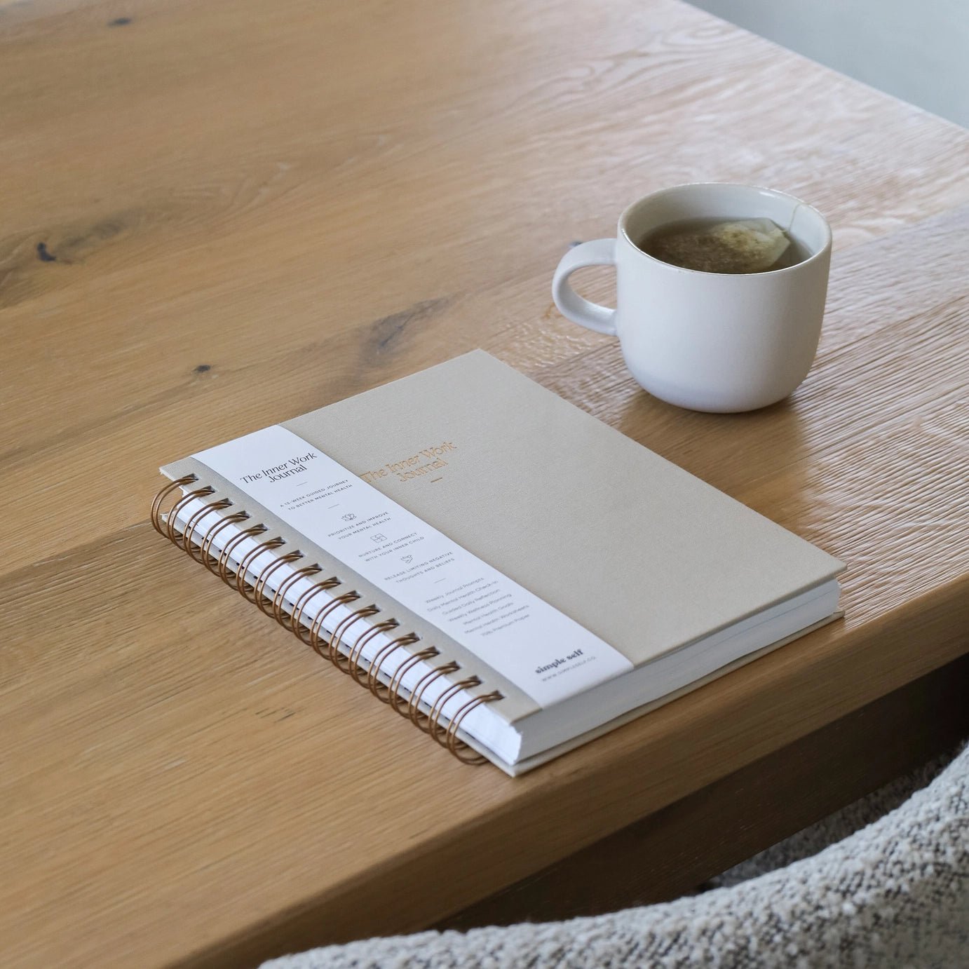 The Inner Work Journal in sand next to a cup of hot tea in a white mug on a wooden table, creating a cozy workspace vibe.